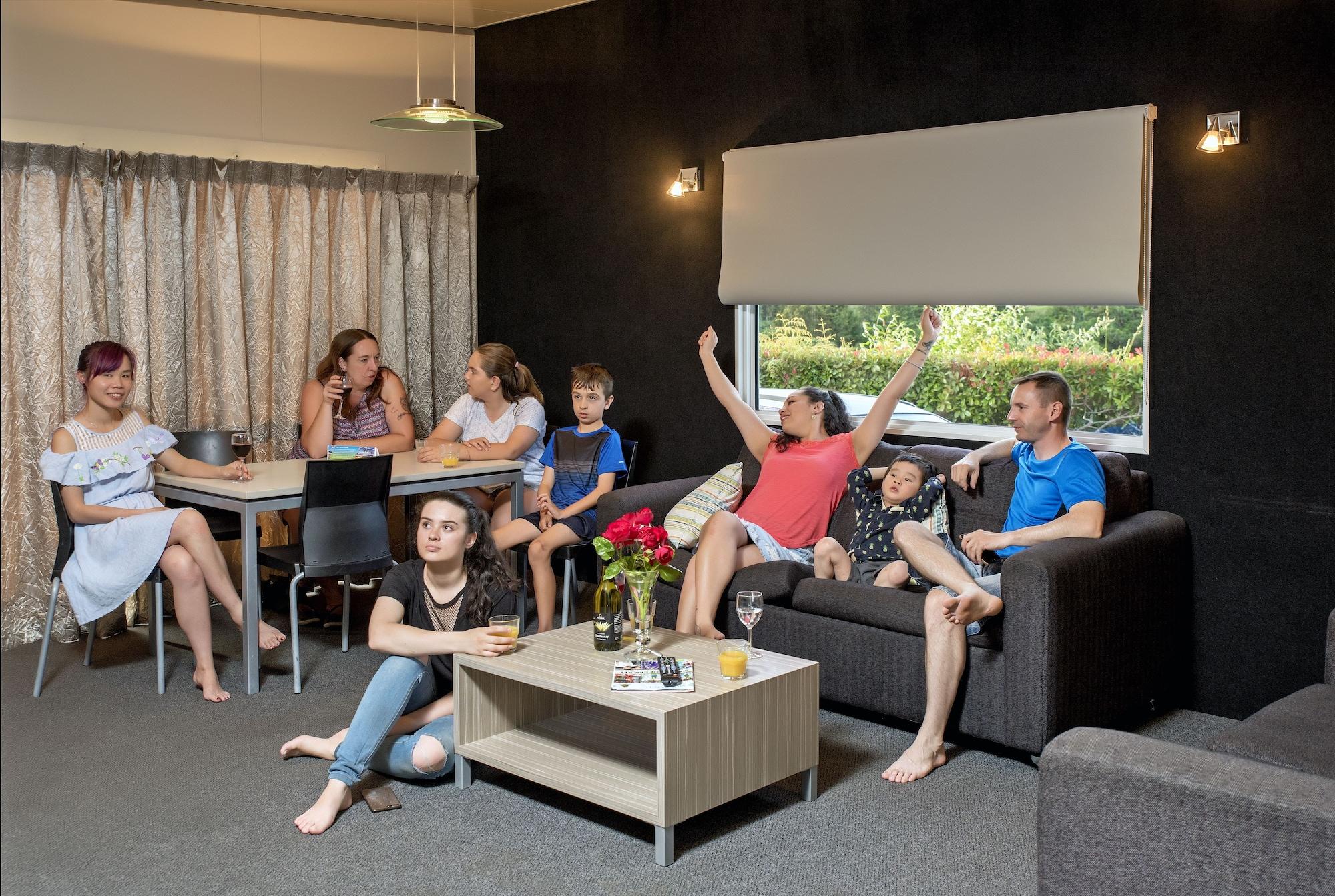 Taupo Debretts Spa Resort Exterior photo The photo shows a group of people gathered in a cozy living room. There are several adults and children interacting with each other. One woman in a red top appears to be celebrating or expressing excitement, while others are engaged in conversation o