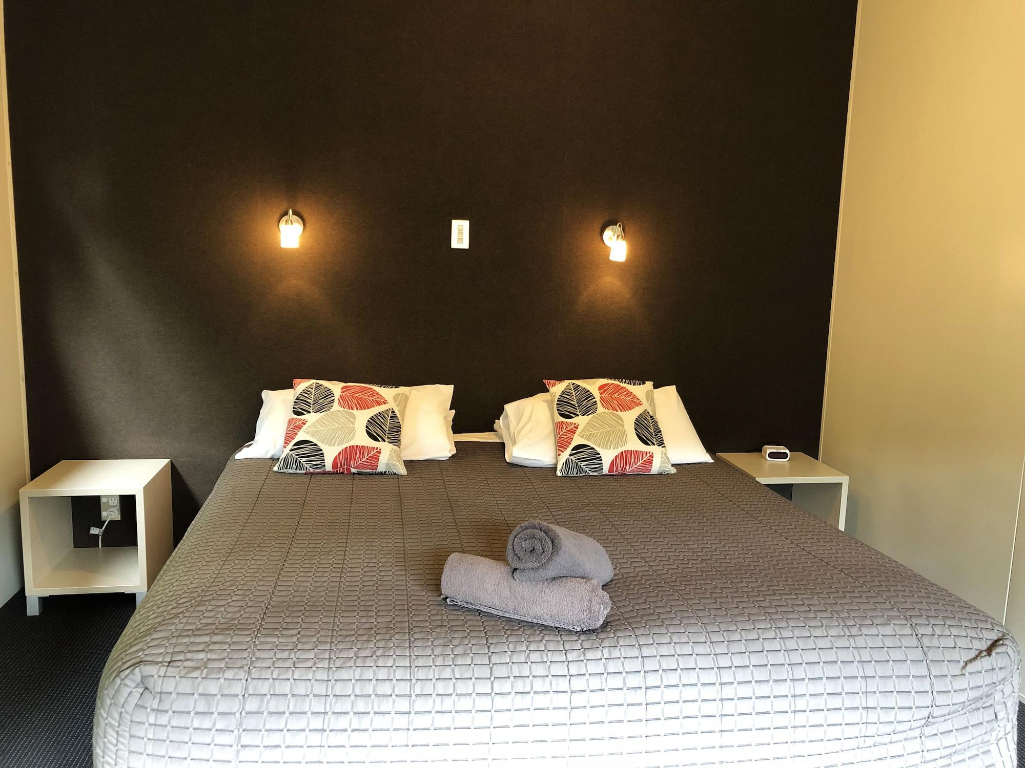 Taupo Debretts Spa Resort Exterior photo The photo shows a bedroom with a large bed that has a gray quilted cover and decorative pillows. The walls behind the bed are painted a dark color, and there are two small wall-mounted lamps providing light. On either side of the bed, there are small